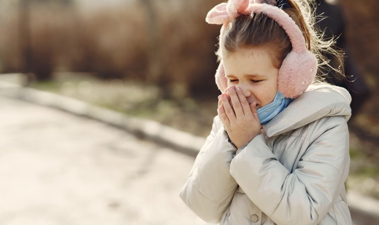 A young girl wearing earmuffs and a coat sneezes outdoors, capturing a moment in cold weather.