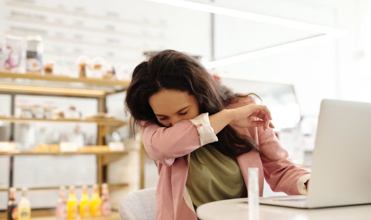 Woman in a cafe sneezing into her elbow while sitting at a table with a laptop.