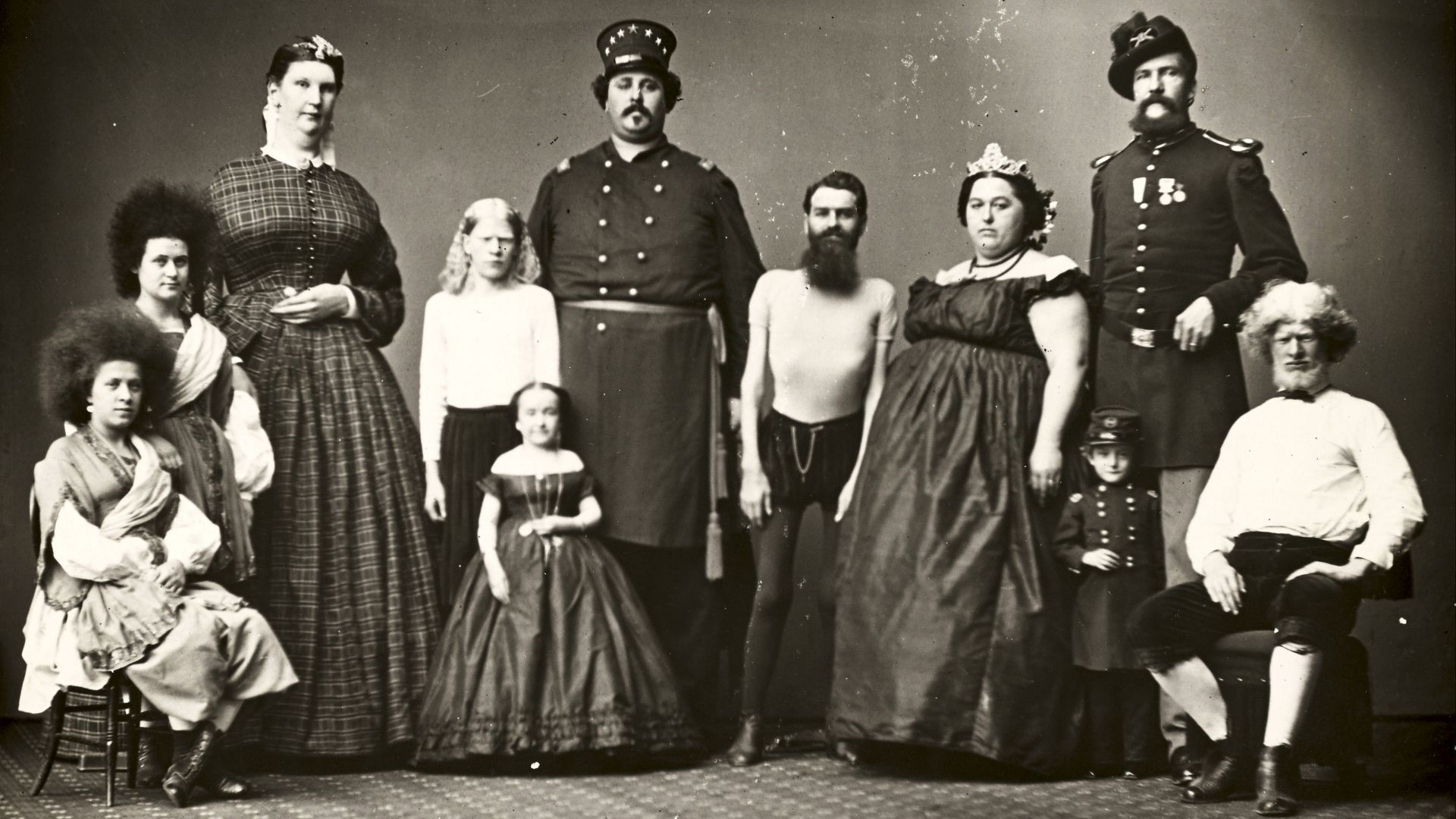 Photograph of group of performers including giants Anna Swan, Noah Orr, and Monsieur Joseph; midgets Lizzie Reid and Edward Newell; albinos Amos Gorhen and Charles Gorhen; Circassian Beauties Zuruby Hannum and Zalumma Agra; and the married skinny man and 