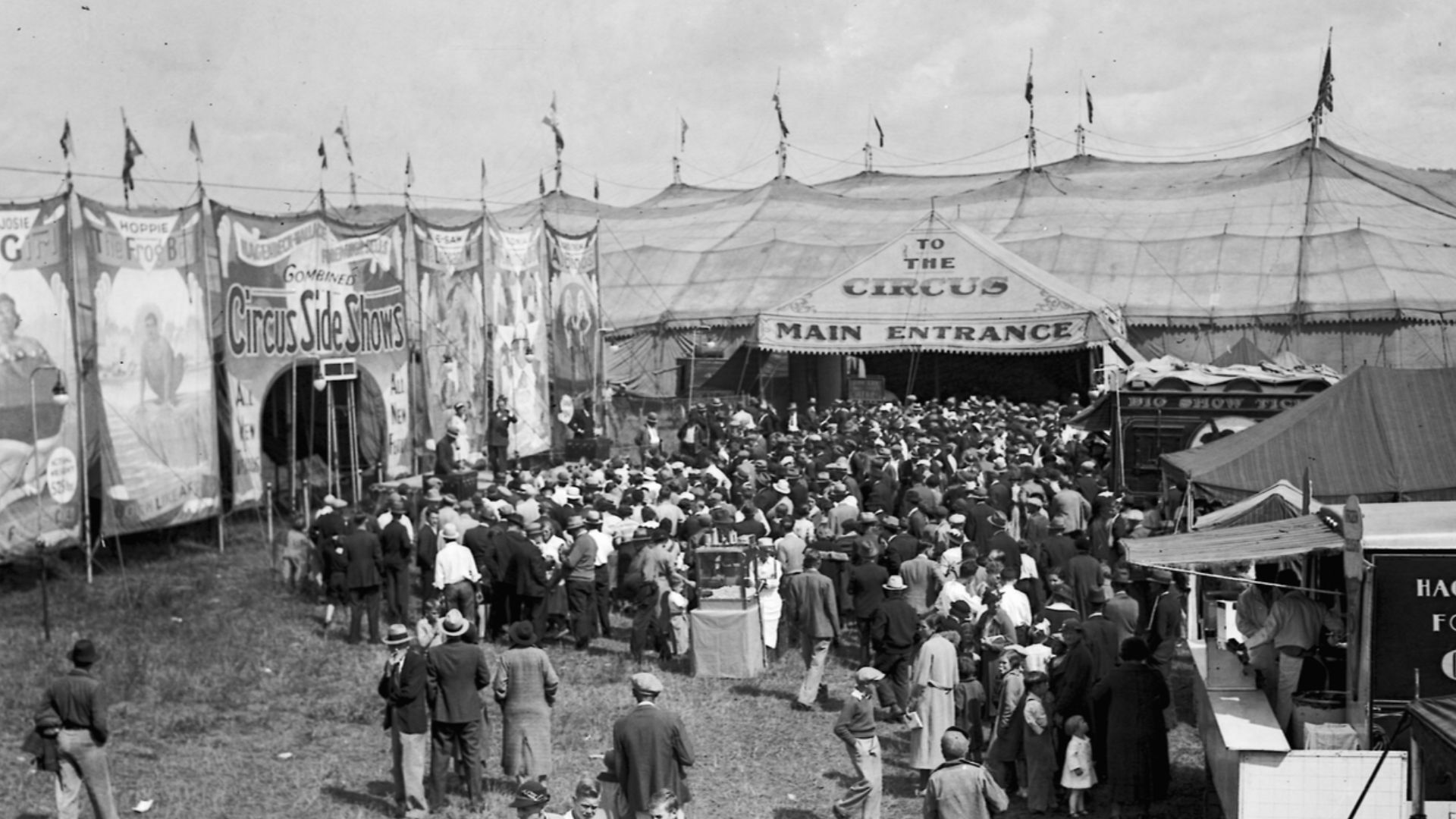 This 1935 photograph shows a crowd gathering on the midway of the Hagenbeck-Wallace Circus, heading towards the entrance marquee tent. On the left is the painted banner line depicting freaks and attractions in the sideshow, an added fee attraction operati
