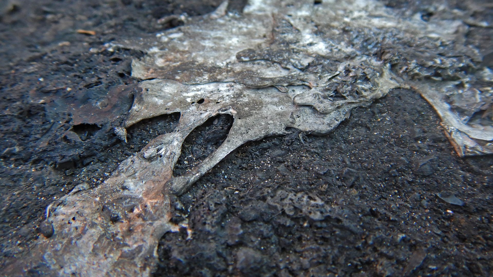 Remnants of melted metals and plastics glued to the tar of a small car park (her after cleaning with the karcher). At this location a car (BMW) was burned a few dozen hours earlier, leaving on the ground molten glass, melted metals and burnt plastic, with