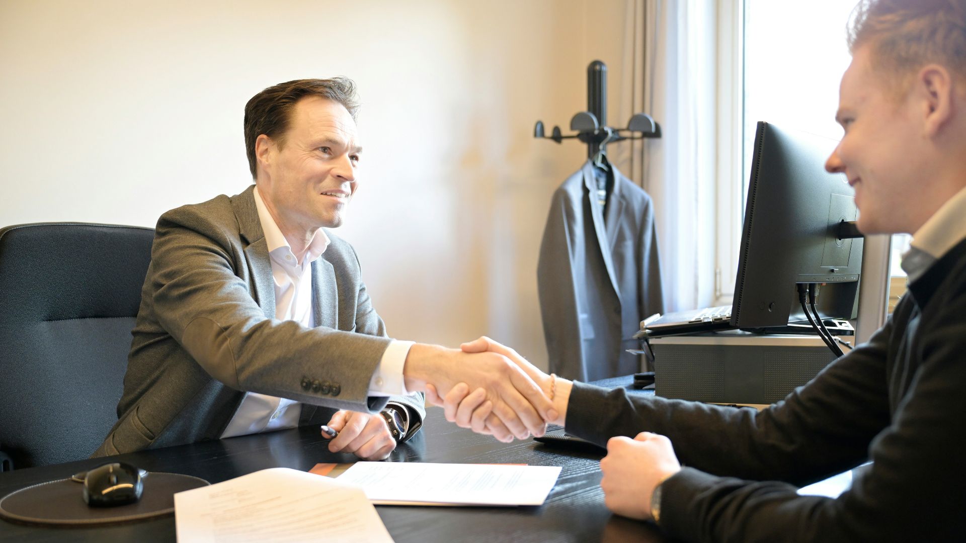 Two smiling men shake hands across an office desk.