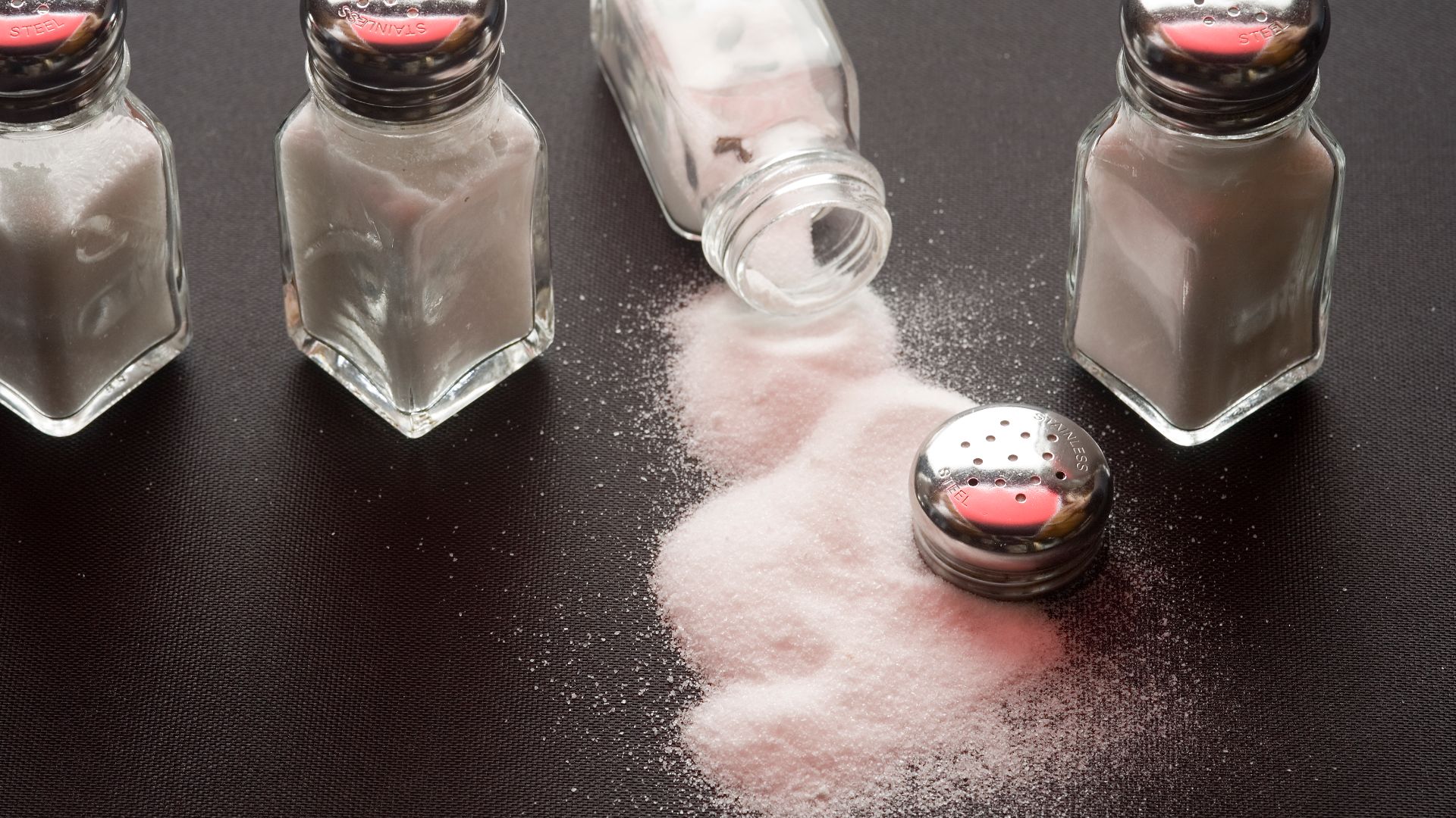 Four salt shakers on a table with salt spilled from one of them.