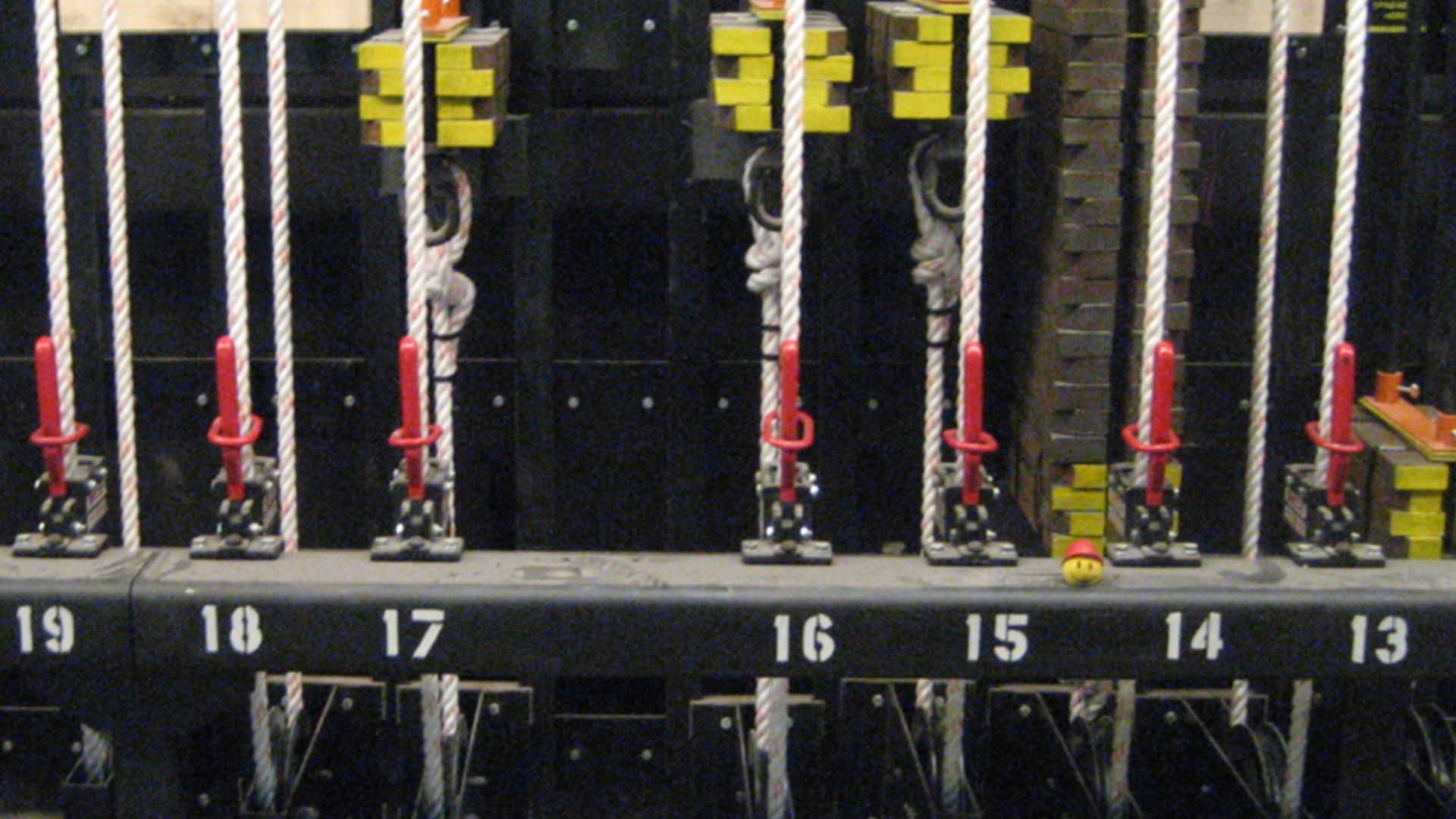 Photograph of the locking rail of a counterweight rigging system.  The steel tube locking rail has rope locks bolted to it with a stenciled number for corresponding the line set.  The operating line passes through the rope lock, around the tension block a