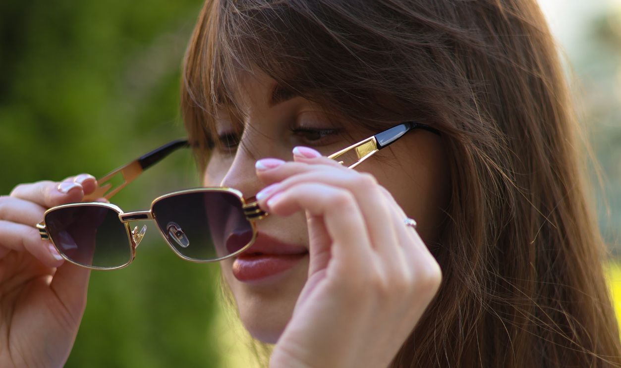Portrait of a young woman adjusting her sunglasses in a sunny park setting.
