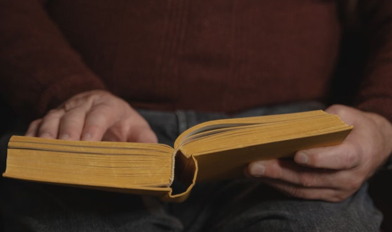 Close-up of a man reading a yellow book while sitting indoors wearing a brown sweater.