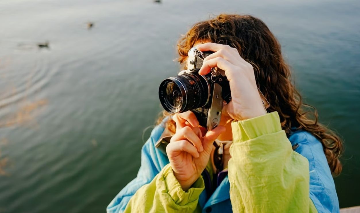 A woman taking photos with a vintage camera by a scenic lake under daylight