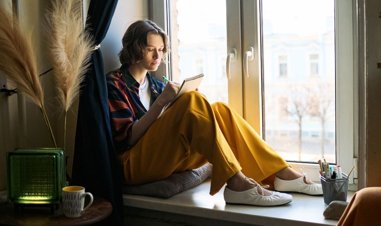 Young adult Caucasian woman sitting on windowsill, writing in notebook and reflecting on ideas. Suitable for mental health, journaling, self care, lifestyle marketing
