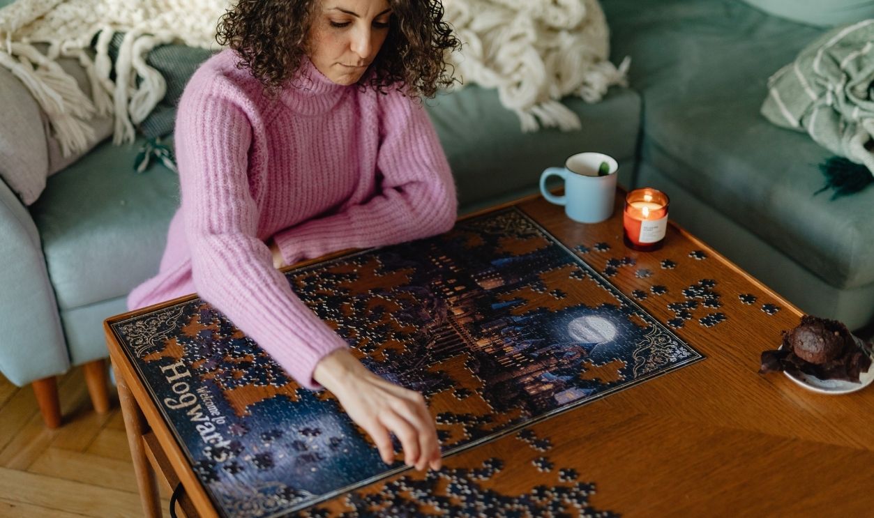A woman in a pink sweater concentrates on solving a puzzle next to a cup and candle