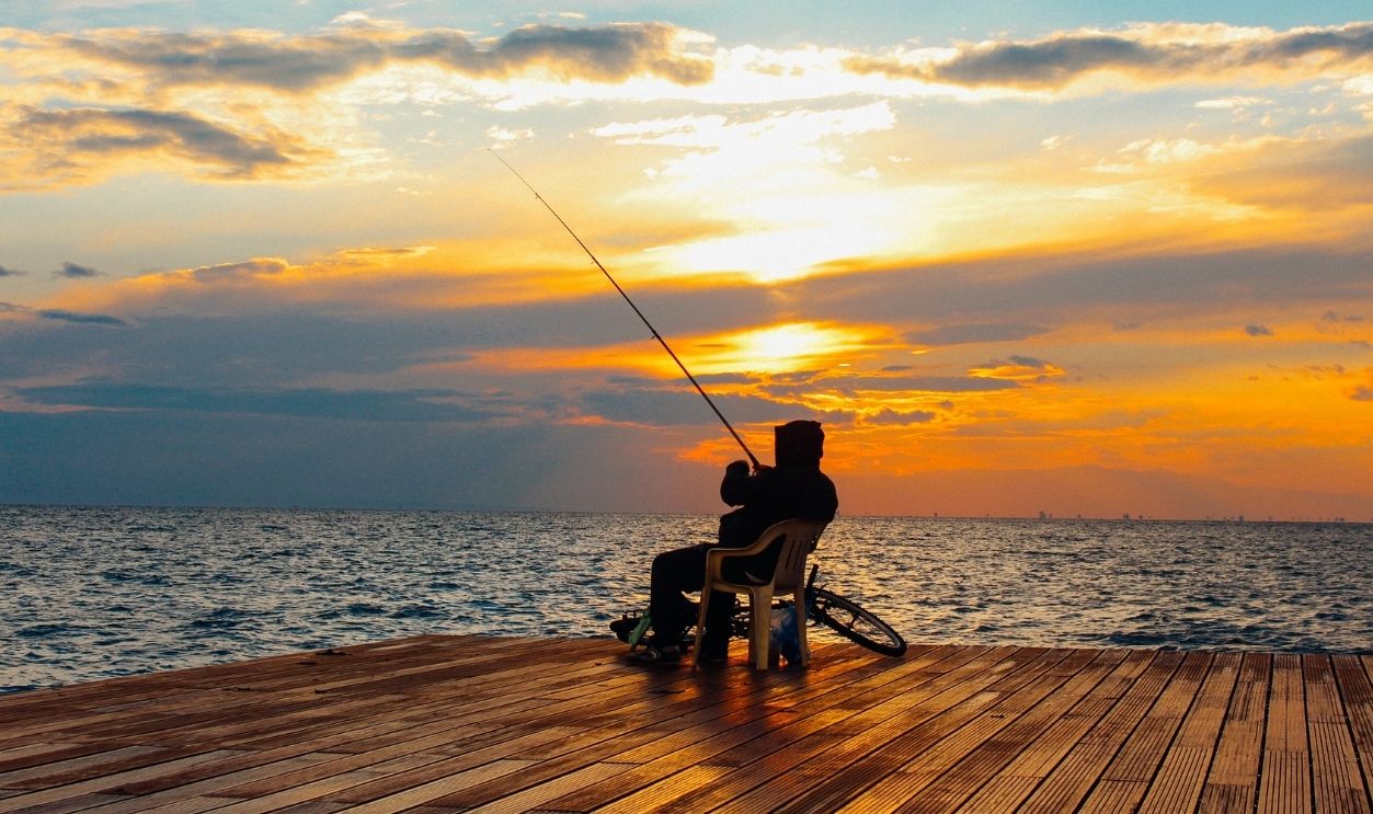 A fisherman sits on a wooden deck, enjoying a peaceful sunset by the ocean, rod in hand