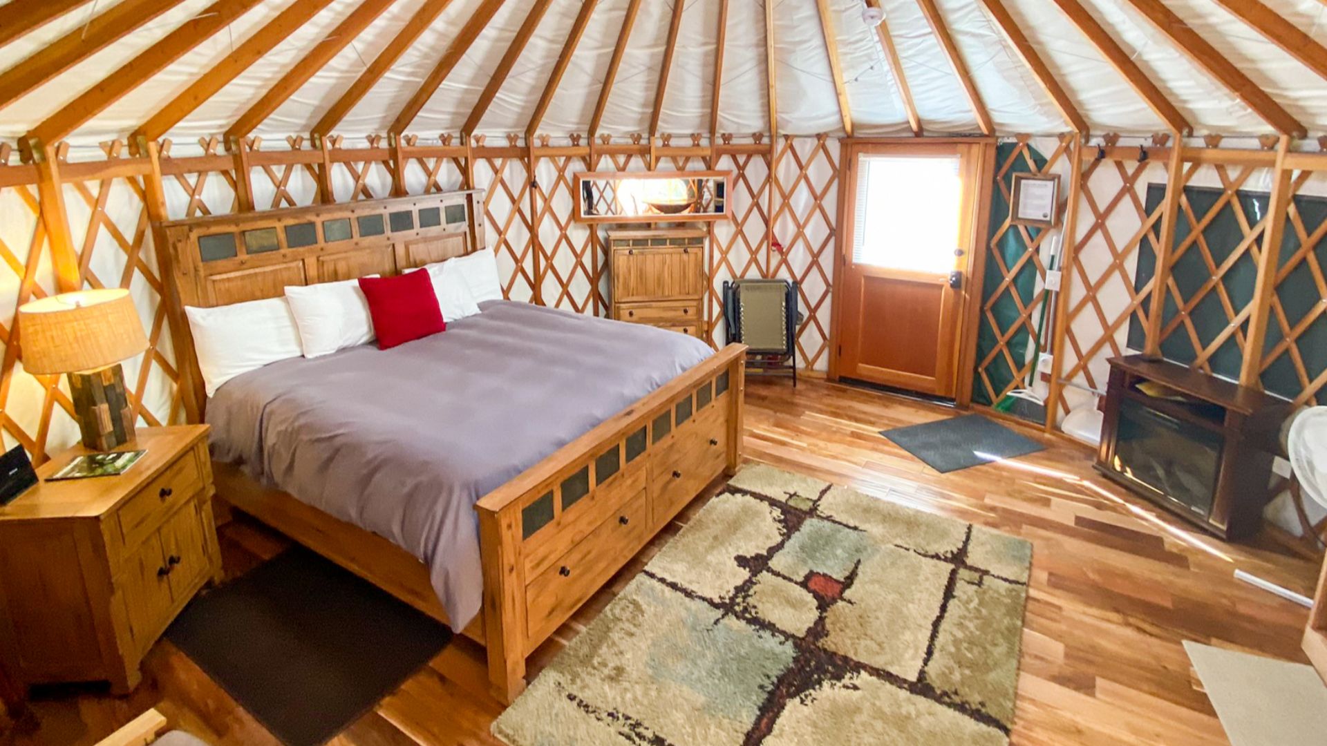 King size bed in a round yurt structure. It has a clear dome on the ceiling, exposed wood beam ceiling and exposed wood lattice wall supports. A couch, dresser, and rug can also be seen.
King size bed in the cozy Willow Glamping Yurt. The windows can be c