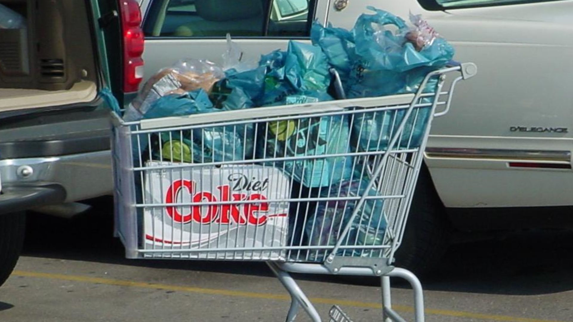 A shopping cart in a Walmart parking lot, filled with plastic grocery bags and a case of Diet Coke cans