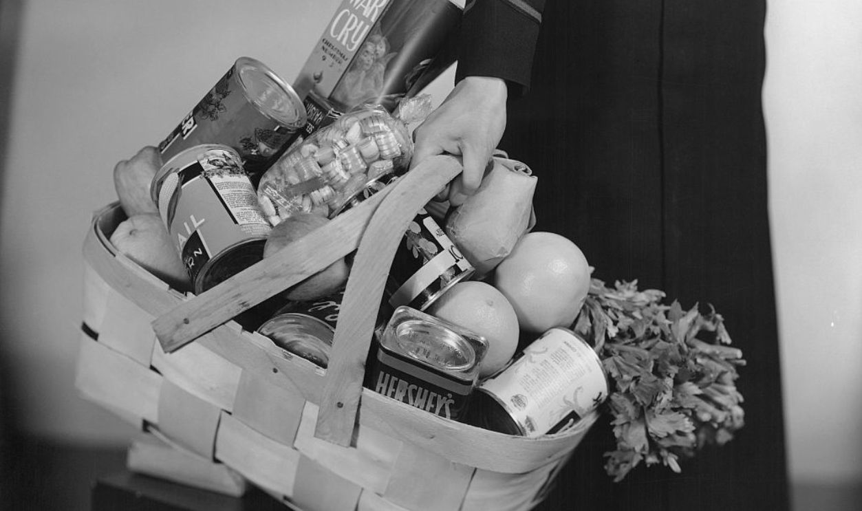 Close-up of a person in black outfit showing their hand on the handles of a woven wooden shopping basket full of groceries.