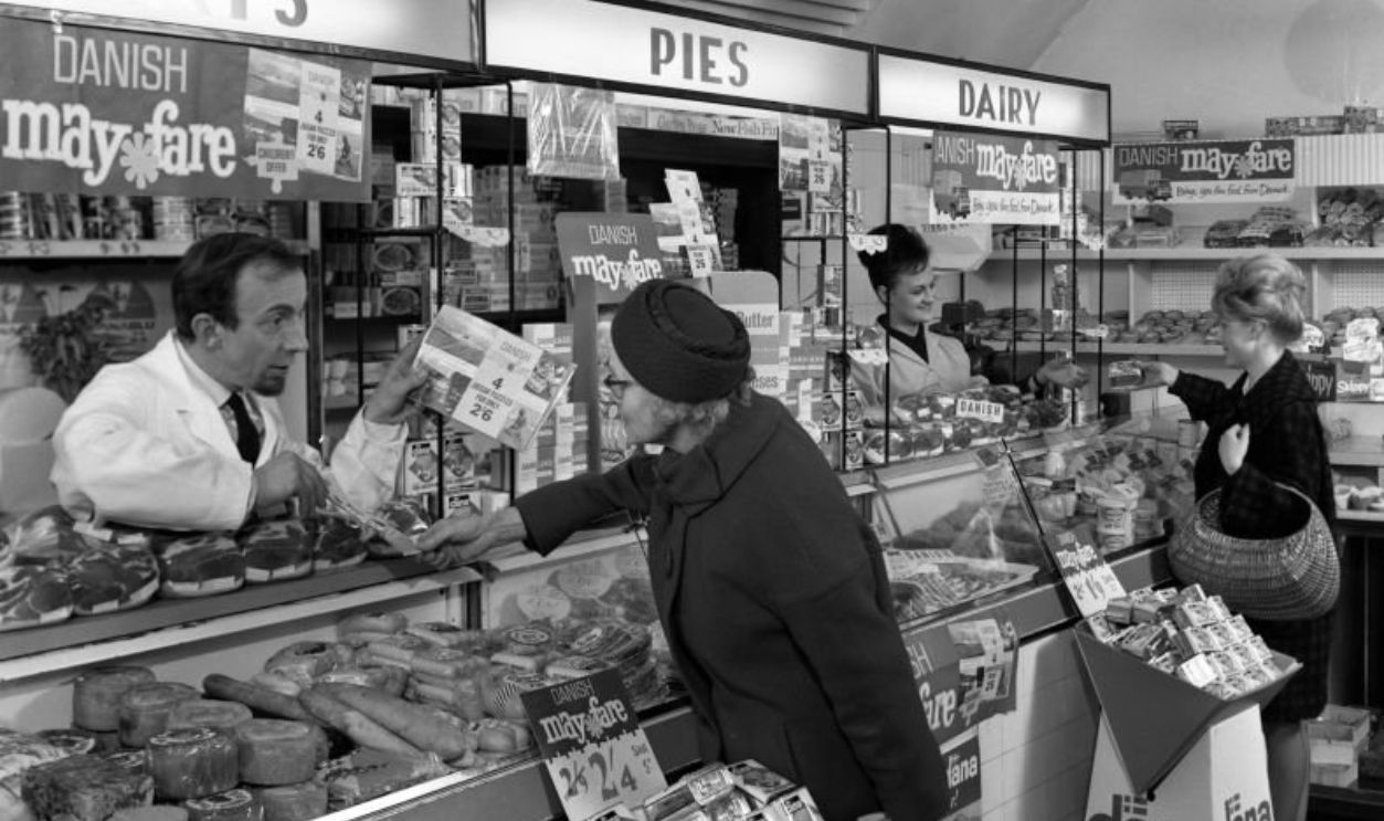 Danish Bacon May Fare shop display, Wath upon Dearne, South Yorkshire, 1964. A shopkeeper in the South Yorkshire town of Wath upon Dearne explains the May offer from Danish Bacon, with an offer of four mini jigsaw puzzles of Danish scenes to a customer.