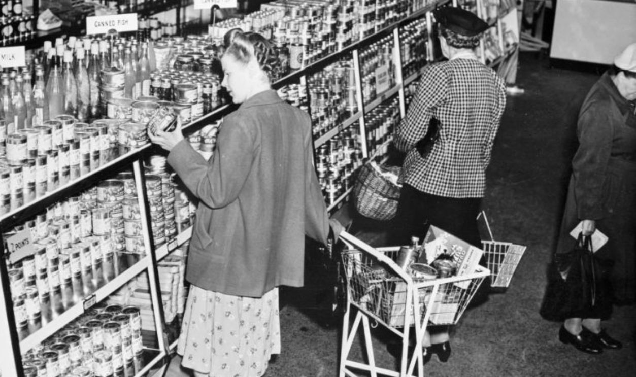 Lyons staff store, Cadby Hall, West Kensington, London. The self service grocery department. Women peruse the neatly stacked shelves of tinned food and bottled drinks.