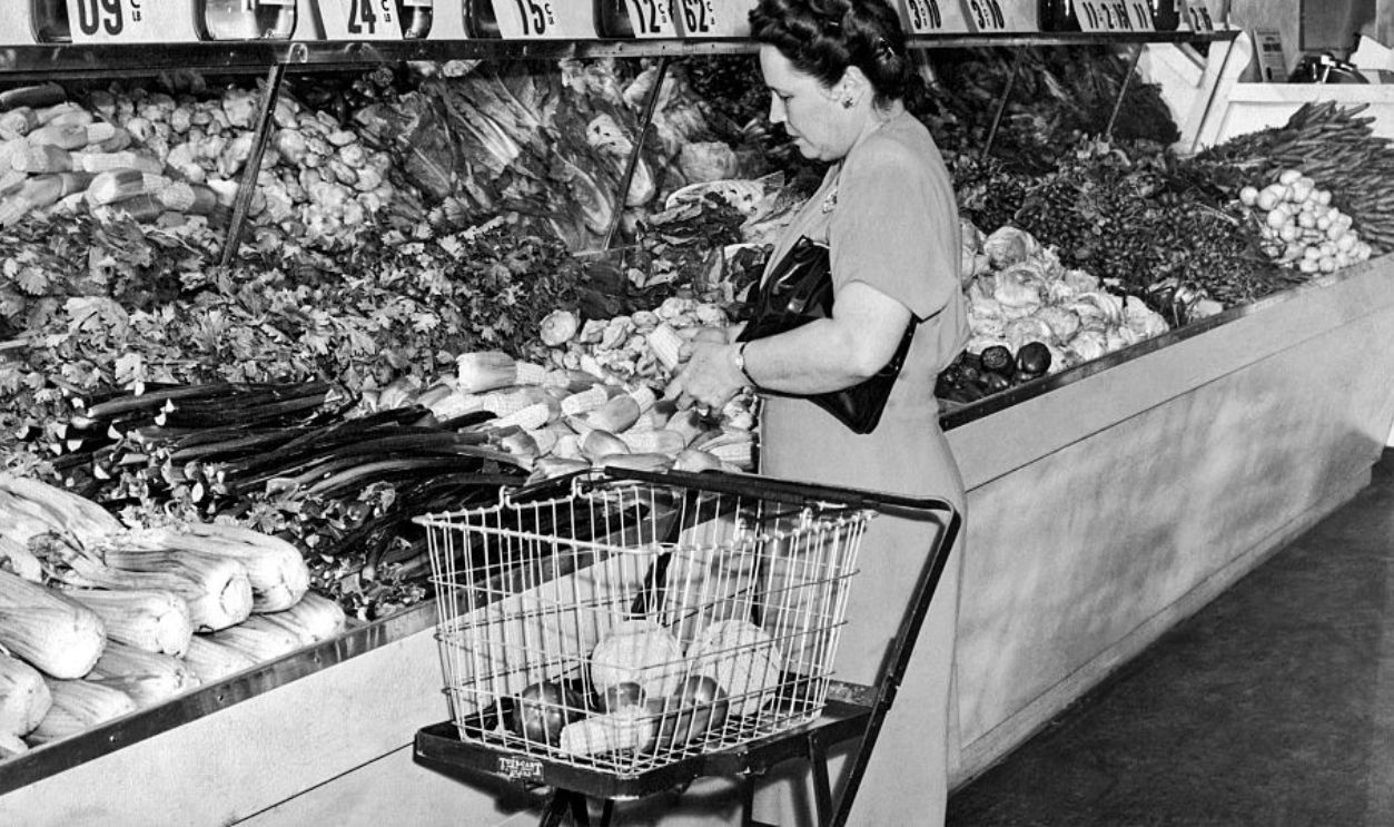 View of a shopper as she selects corn in the vegetable department on an unspecified grocery store, circa 1940. Beside her is a basket on a Tote-Cart brand foldable shopping cart. 