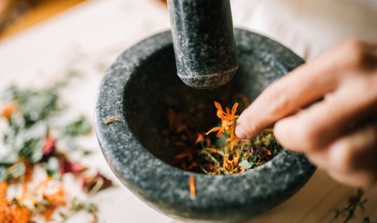 A hand crushes vibrant herbs in a stone mortar, blending natural ingredients
