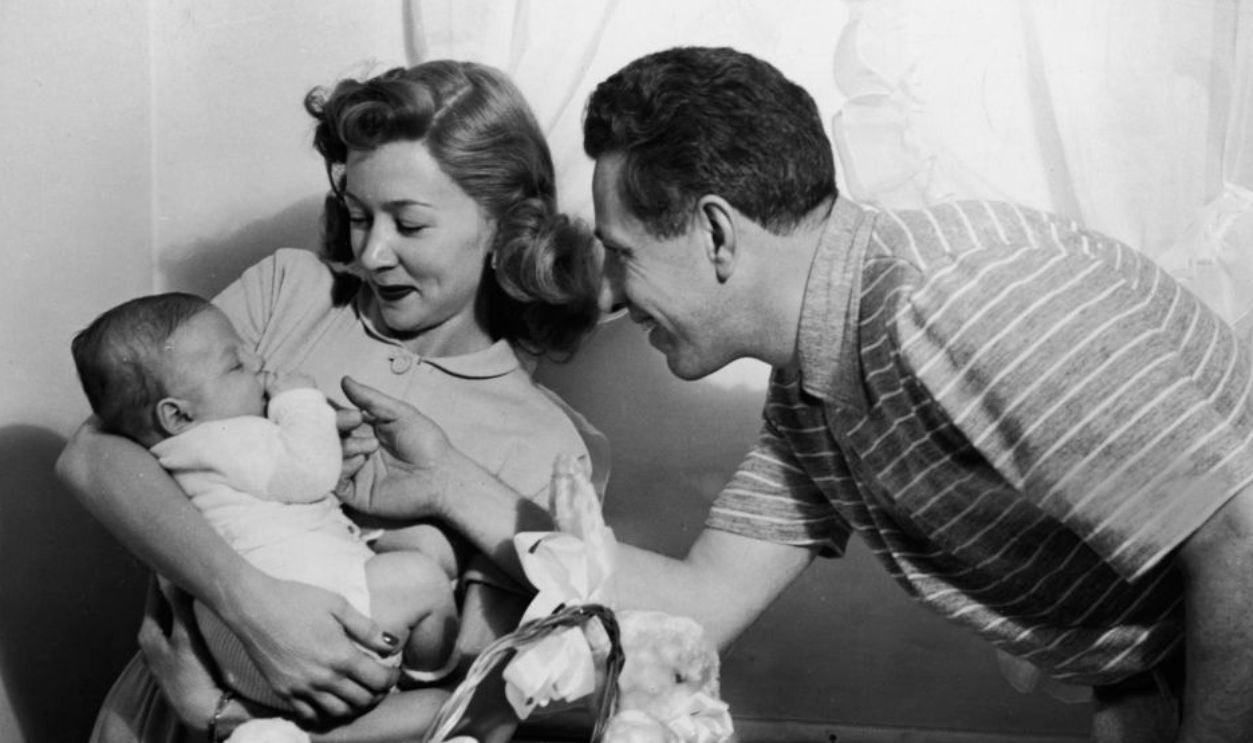 American film director Nicholas Ray (1911 - 1979) and his second wife, American actress Gloria Grahame (1923 - 1981), smile at their three-and-a-half-month-old son, Timothy Nicholas, April 1949. An Easter basket and stuffed animals are on top of a diaper changing station. 