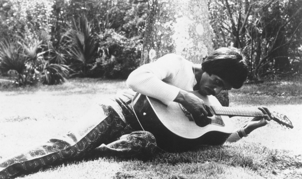 Rock and roll musician Ike Turner poses for a portrait laying on some grass with an acoustic guitar in circa 1971