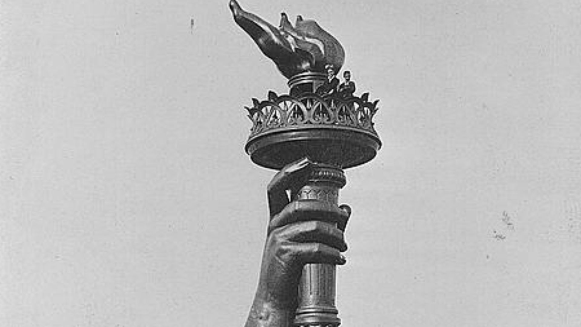 Photograph shows the torch and part of the arm of the Statue of Liberty, on display at the 1876 Centennial Exhibition in Philadelphia. Information booth at base of arm and two persons seen at railing below flame of torch.