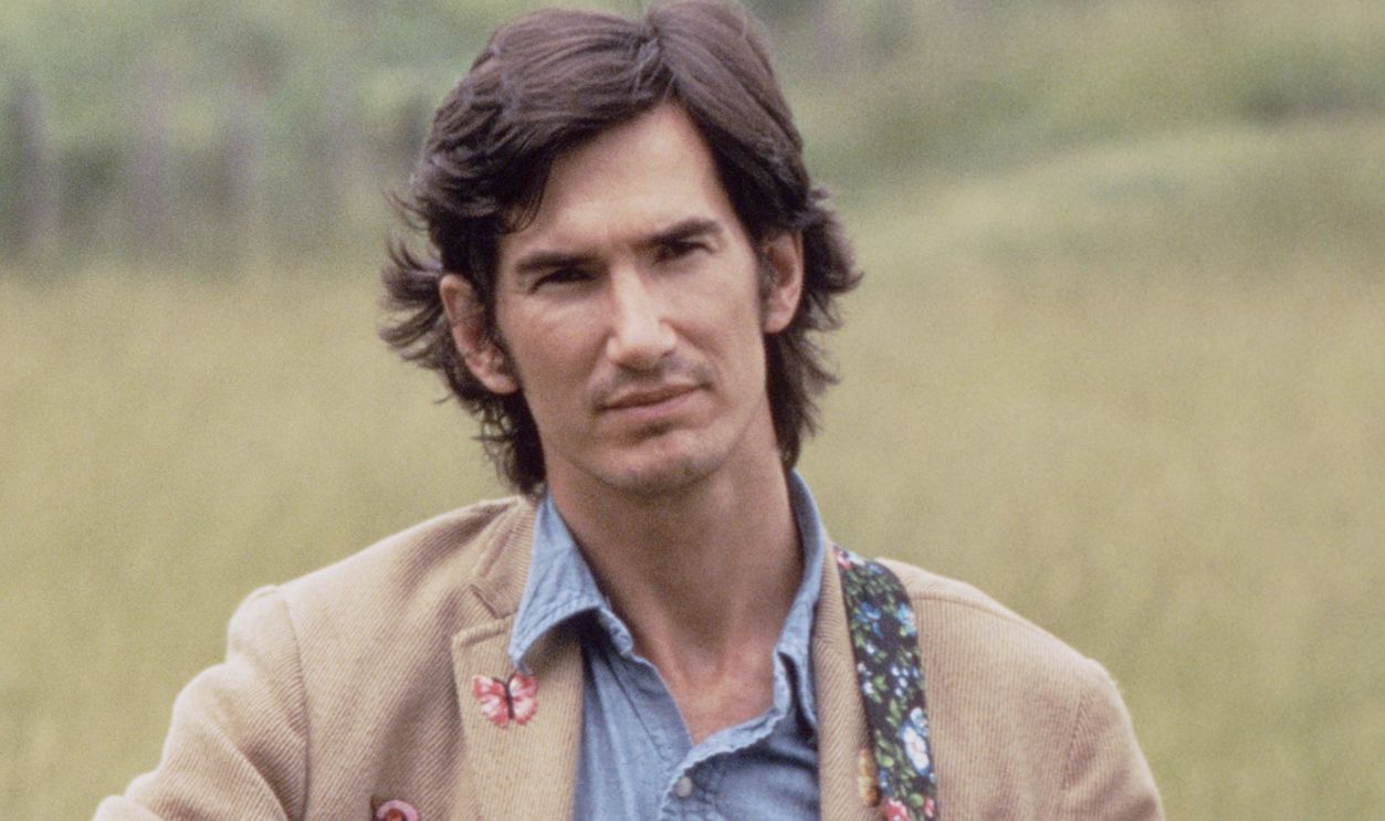 Singer, songwriter, musician, Townes Van Zandt shot waist up, standing in a field with winding road behind him, holding an acoustic guitar wearing beige sport coat, button down shirt and blue jeans. Atlanta, Georgia, circa 1972. Photo is part of the Nashville Music Collection.