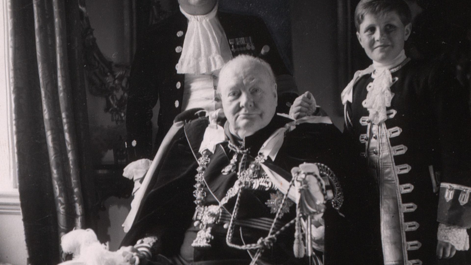 Photograph shows Winston Churchill, full-length portrait, seated, facing front, with his son and grandson standing behind him, wearing coronation robes, at the coronation of Queen Elizabeth II