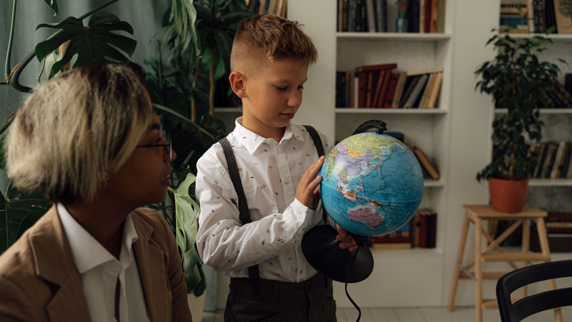 A young boy studying geography indoors with a globe and teacher nearby.