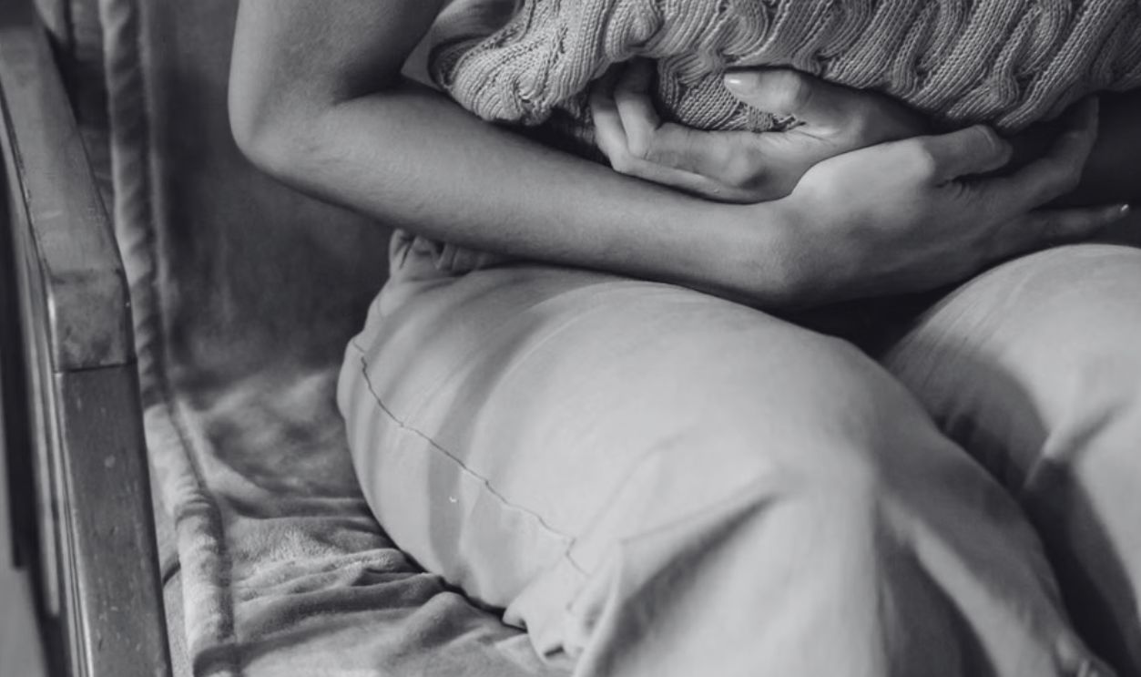 A woman sitting indoors holding a pillow against her stomach, indicating discomfort or pain