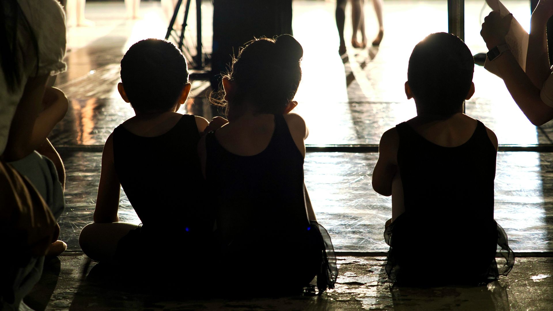 Three children in black leotards sitting on a stage floor during a rehearsal