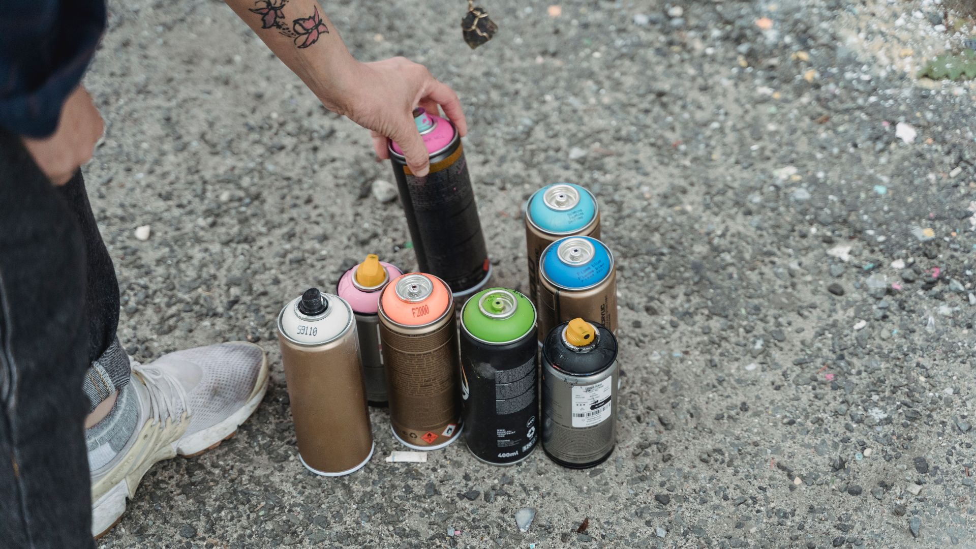 Crop anonymous person in sneakers with tattoo and heap of multicolored spray paint cans on ground standing on street in city