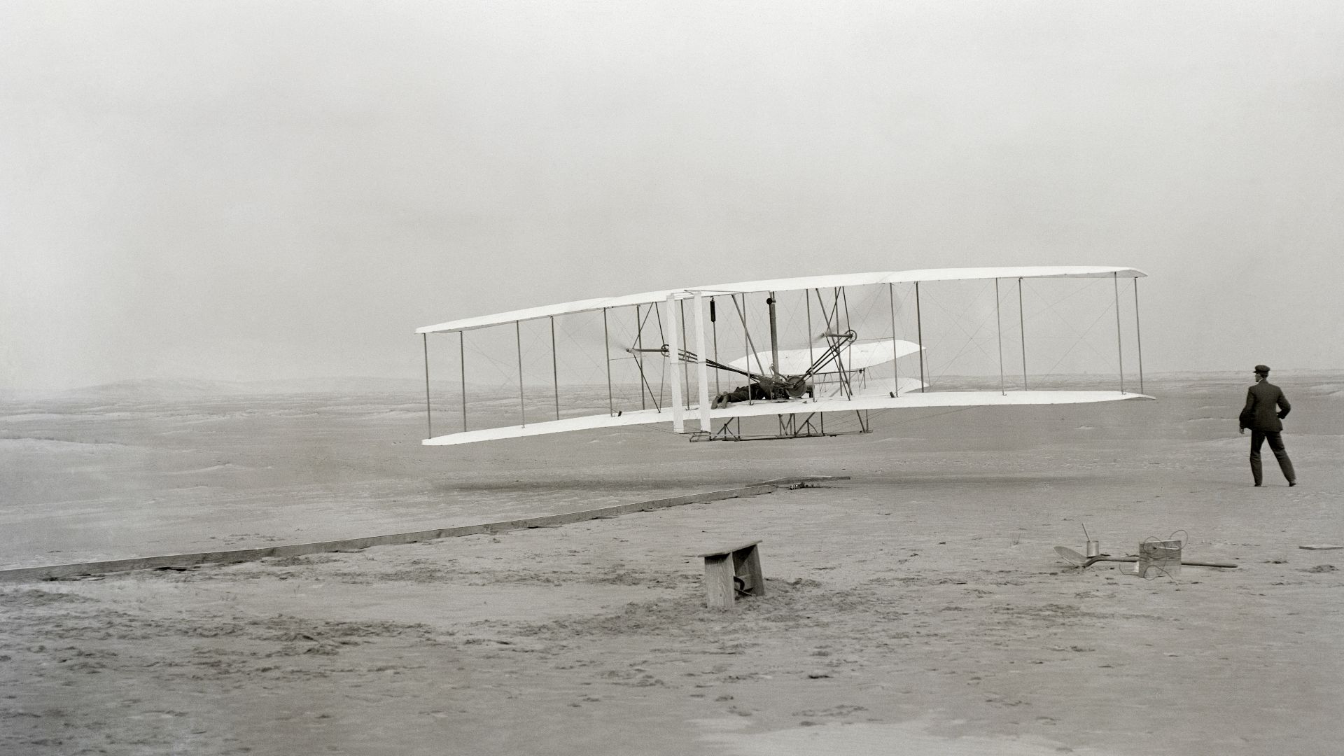 The first powered, controlled, sustained airplane flight in history. Orville Wright, age 32, is at the controls of the machine, lying prone on the lower wing with hips in the cradle which operated the wing-warping mechanism. His brother, Wilbur Wright, ag