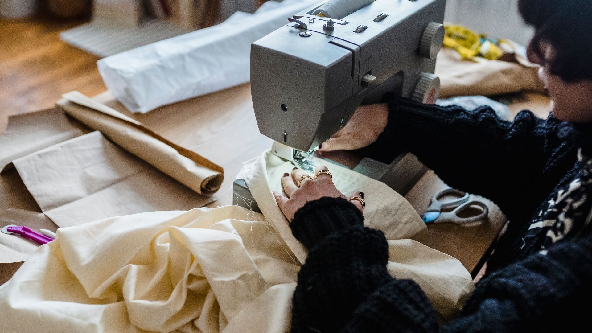 From above of crop female sewer in stylish warm sweater stitching fabric with sewing machine while working in traditional atelier