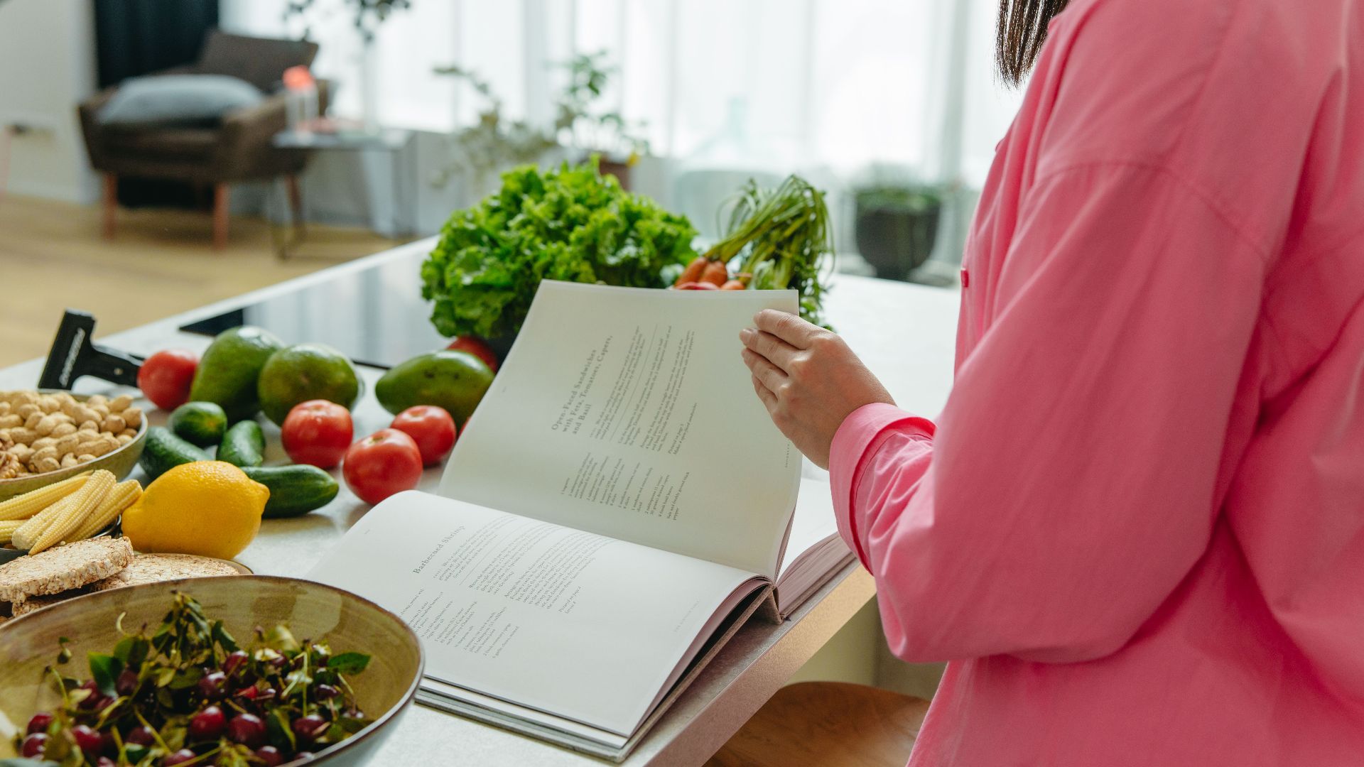 A woman in a kitchen flipping through a cookbook surrounded by fresh vegetables.