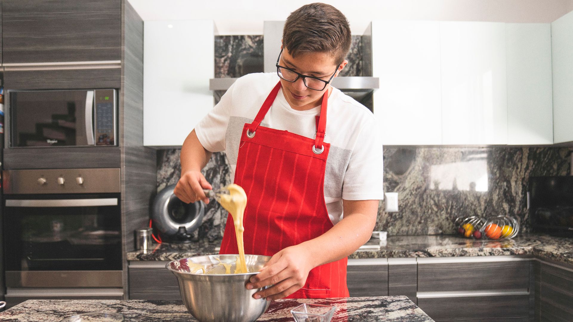 Teenager in a modern kitchen wearing a red apron while whisking ingredients in a stainless steel bowl.