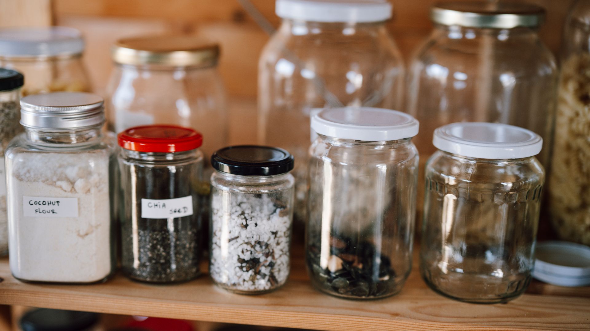 Shelving in a kitchen pantry shows various jars and bottles storing dry food items. The organized setup includes grains, seeds, and snacks. Labels help identify each container's contents.