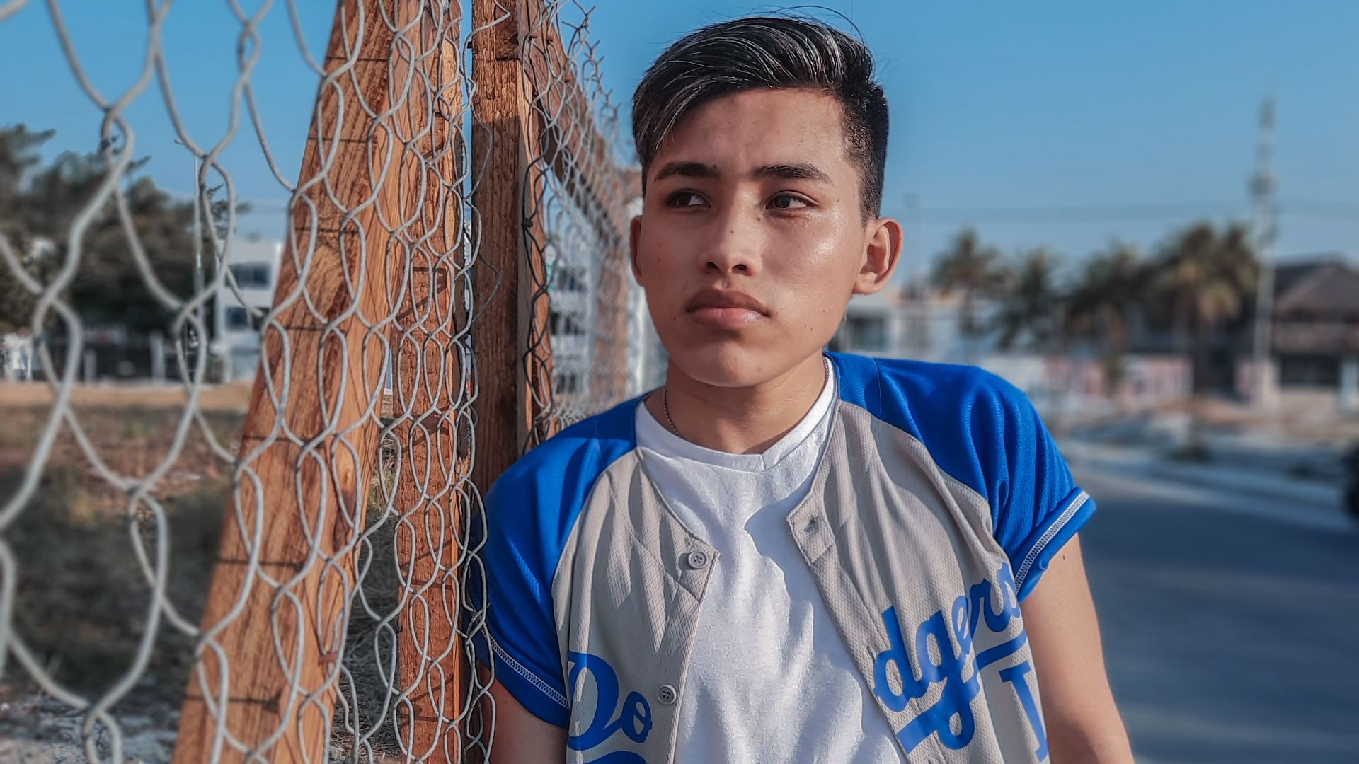 Teenage boy in casual attire leans on a fence outdoors on a sunny day, conveying urban style and confidence.
