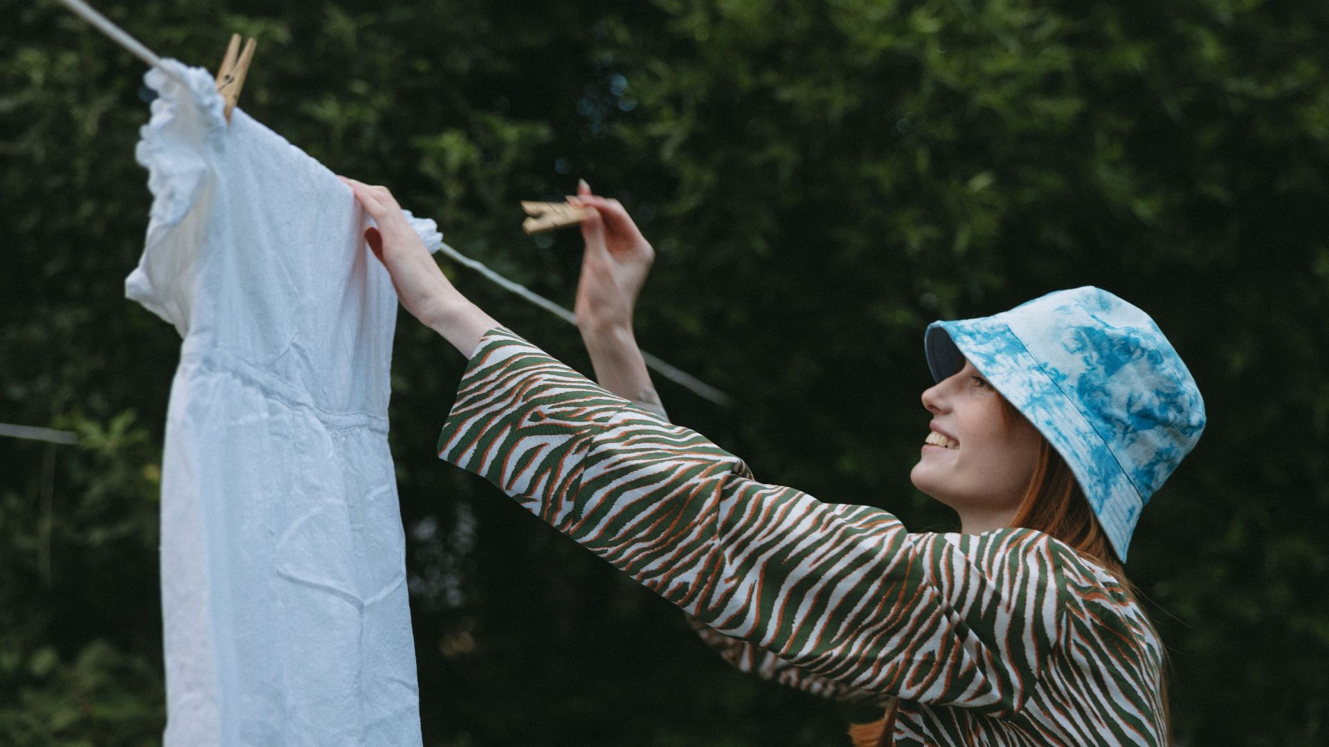 Smiling woman hanging white dress on clothesline in garden, enjoying a sunny day.