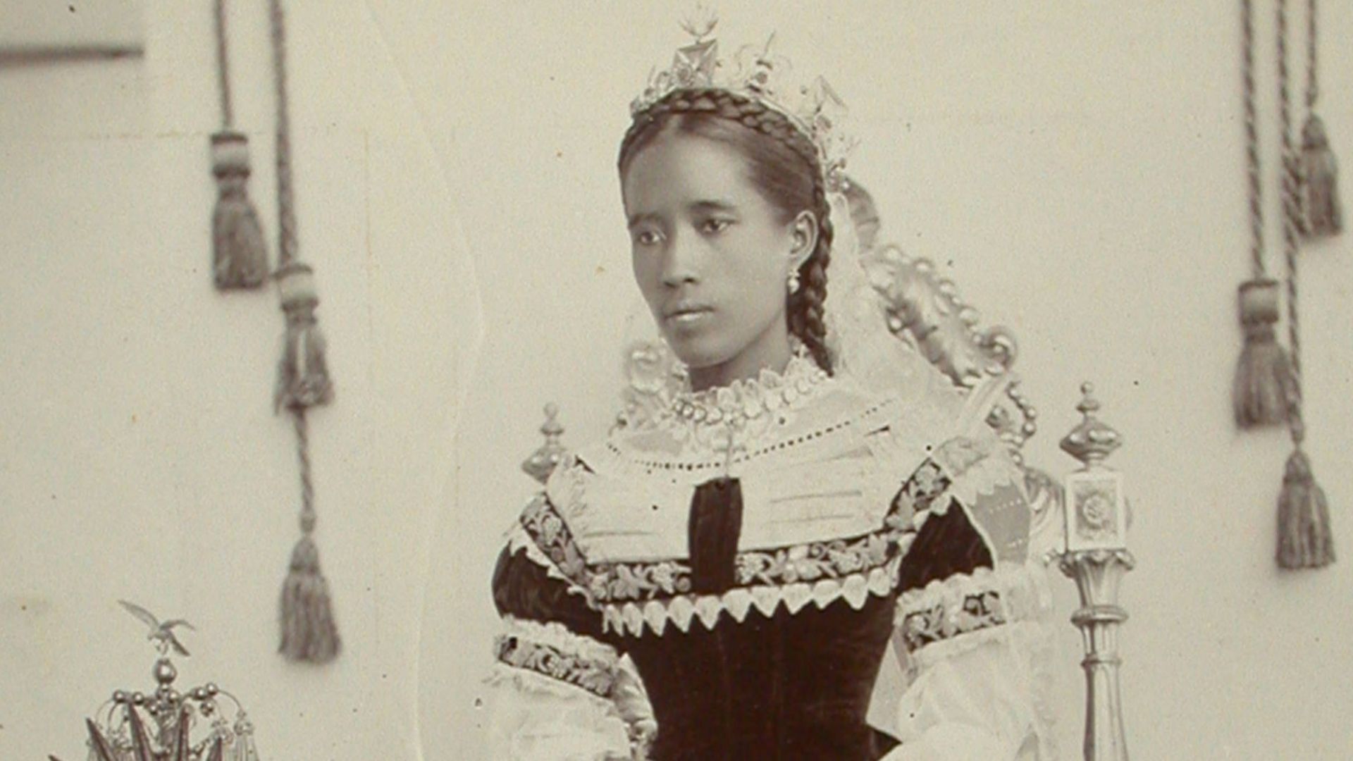 Queen Ranavalona III standing beside a throne table which her crown is lying on. She is wearing a beautiful royal gown which has some embroidery on it. Her hair is braided.