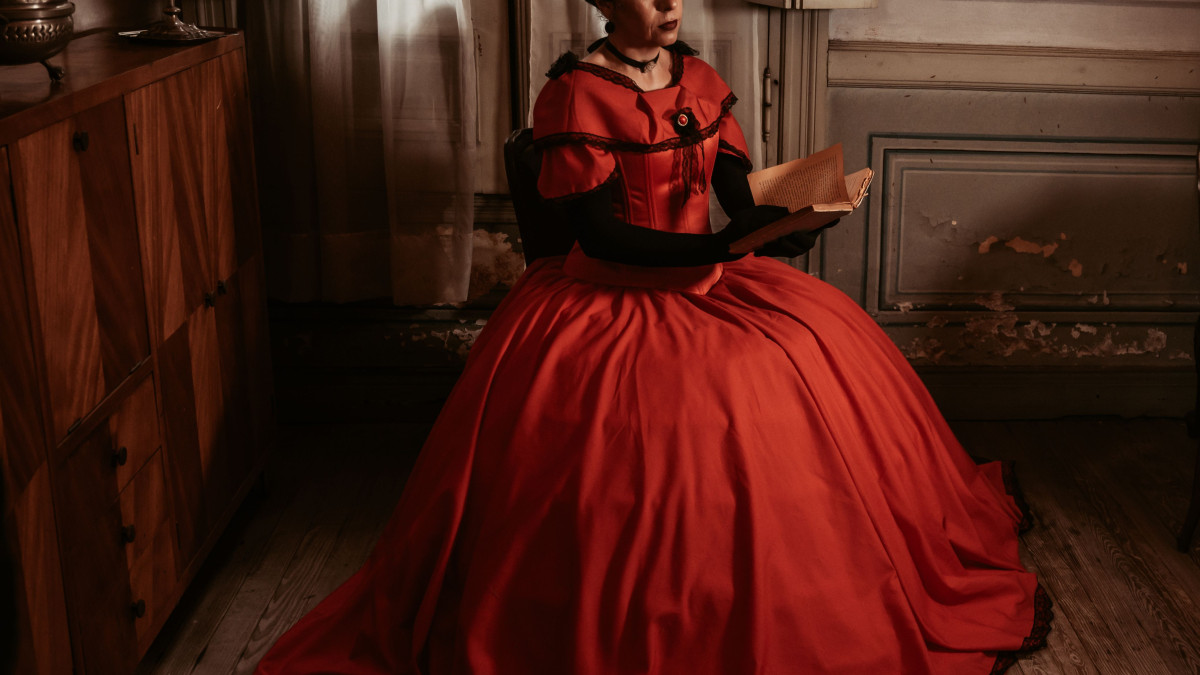 A Victorian-era woman sits in a dimly lit room, elegantly dressed in a red gown, absorbed in reading.