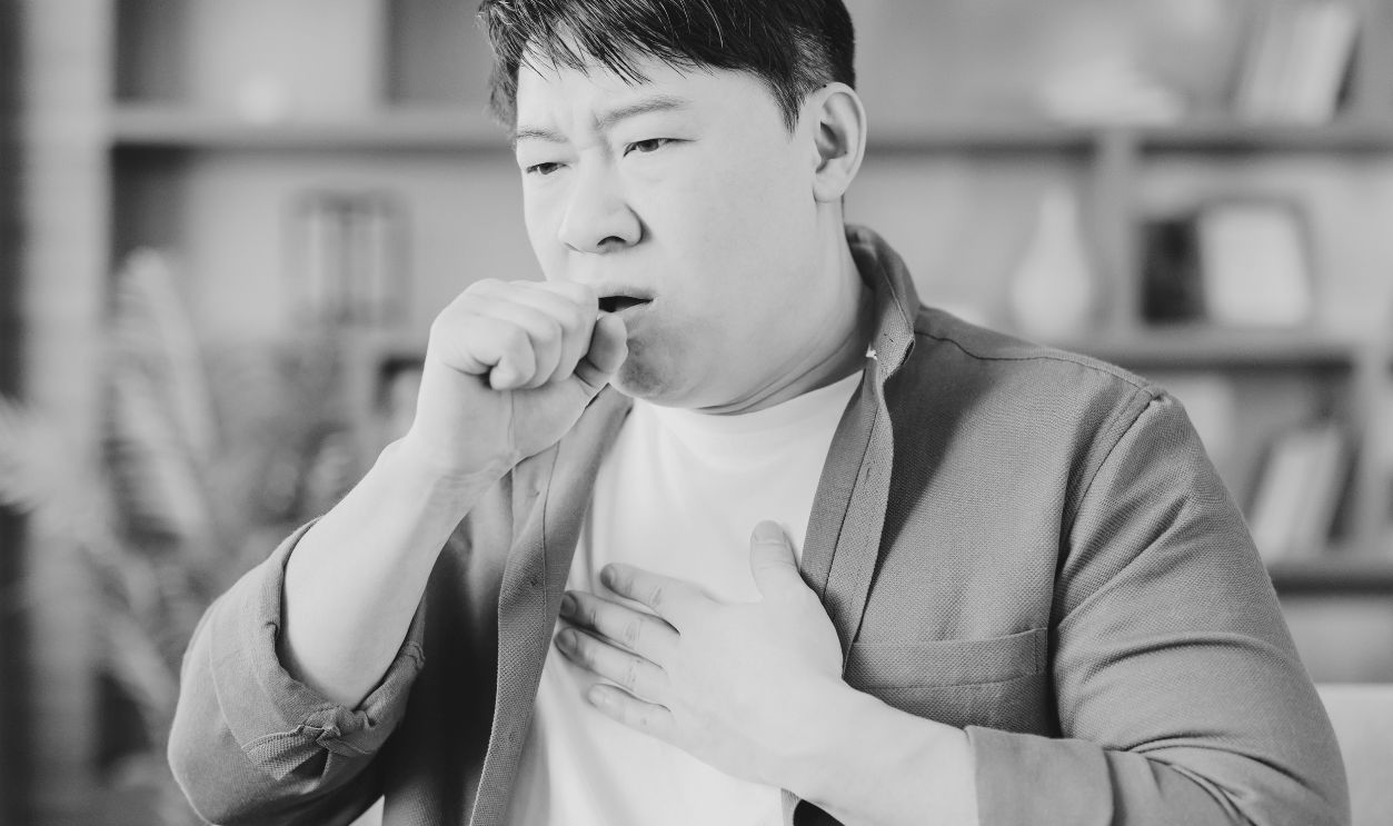 Asian man at home clutching chest and covering his mouth while coughing, showing flu-like symptoms and discomfort as he rests in a living room during illness and recovery