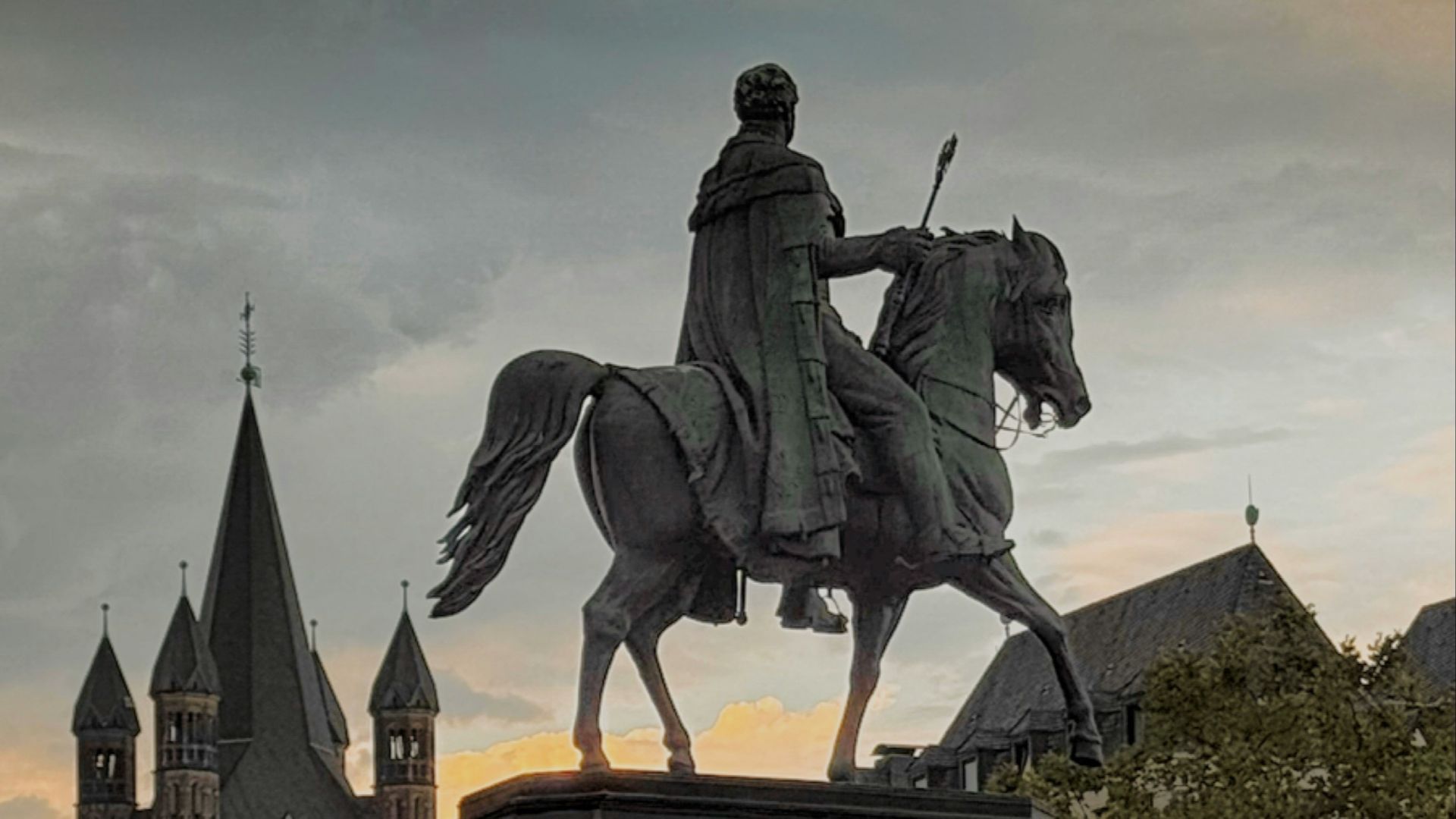 A bronze equestrian statue on a stone pedestal in a city square at dusk