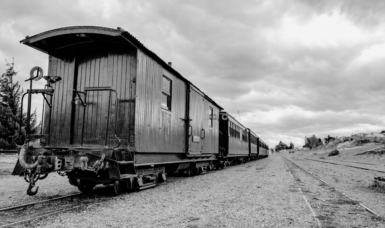 A rustic wooden train car sits on railway tracks under a cloudy sky, evoking a vintage feel