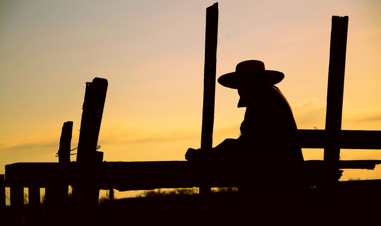 Silhouette of a man in a hat standing by a wooden fence during sunset. Dramatic sky enhances the scene