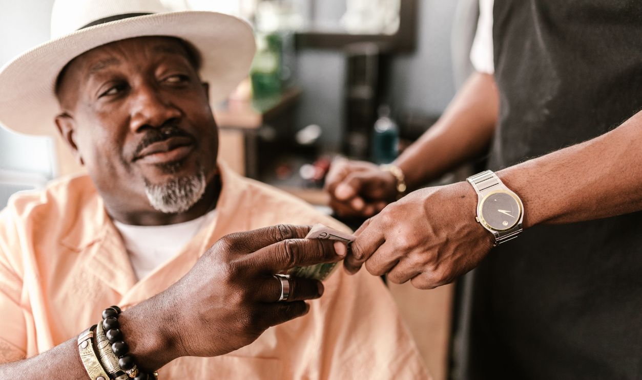 Senior man with a hat giving a tip to a young barber indoors, wearing bracelets and a watch.