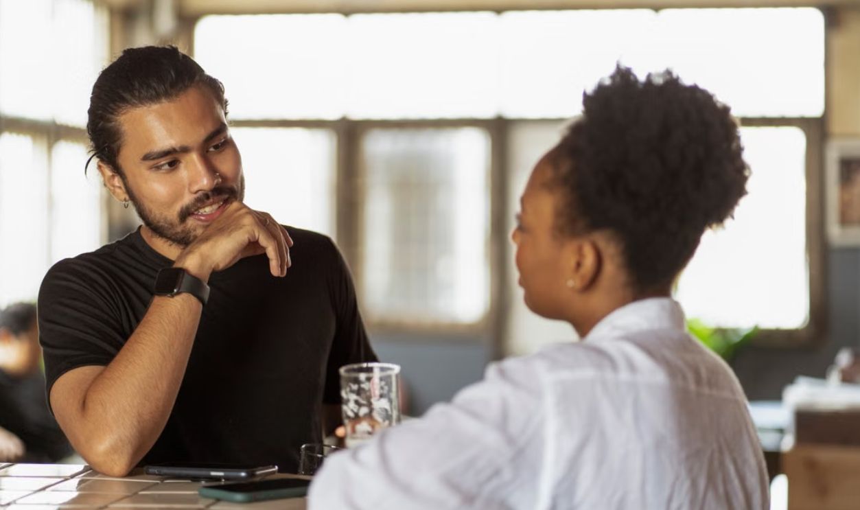Two adults conversing at a table indoors, with natural light through windows