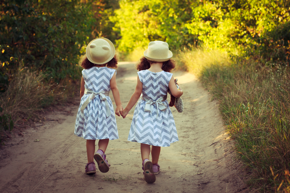 Two cute curly kids in a beautiful dress and straw summer hats walking away on forest or park road.