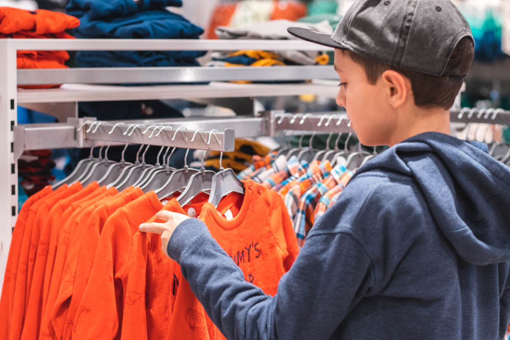 Young boy choosing clothes in the shop by himself, putting on new looks, checking for style