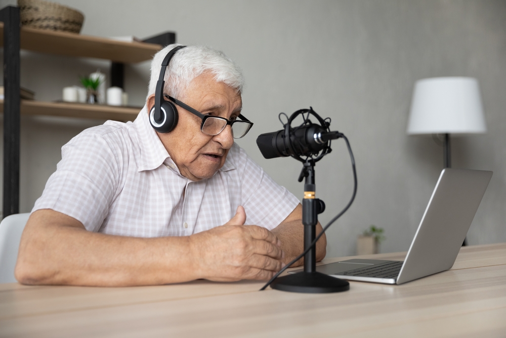 Focused elderly retired man is wearing wireless headphones and looking at computer screen.