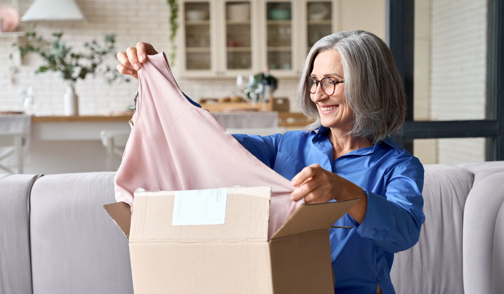 Happy senior aged woman in blue shirt opening box with ordered clothes