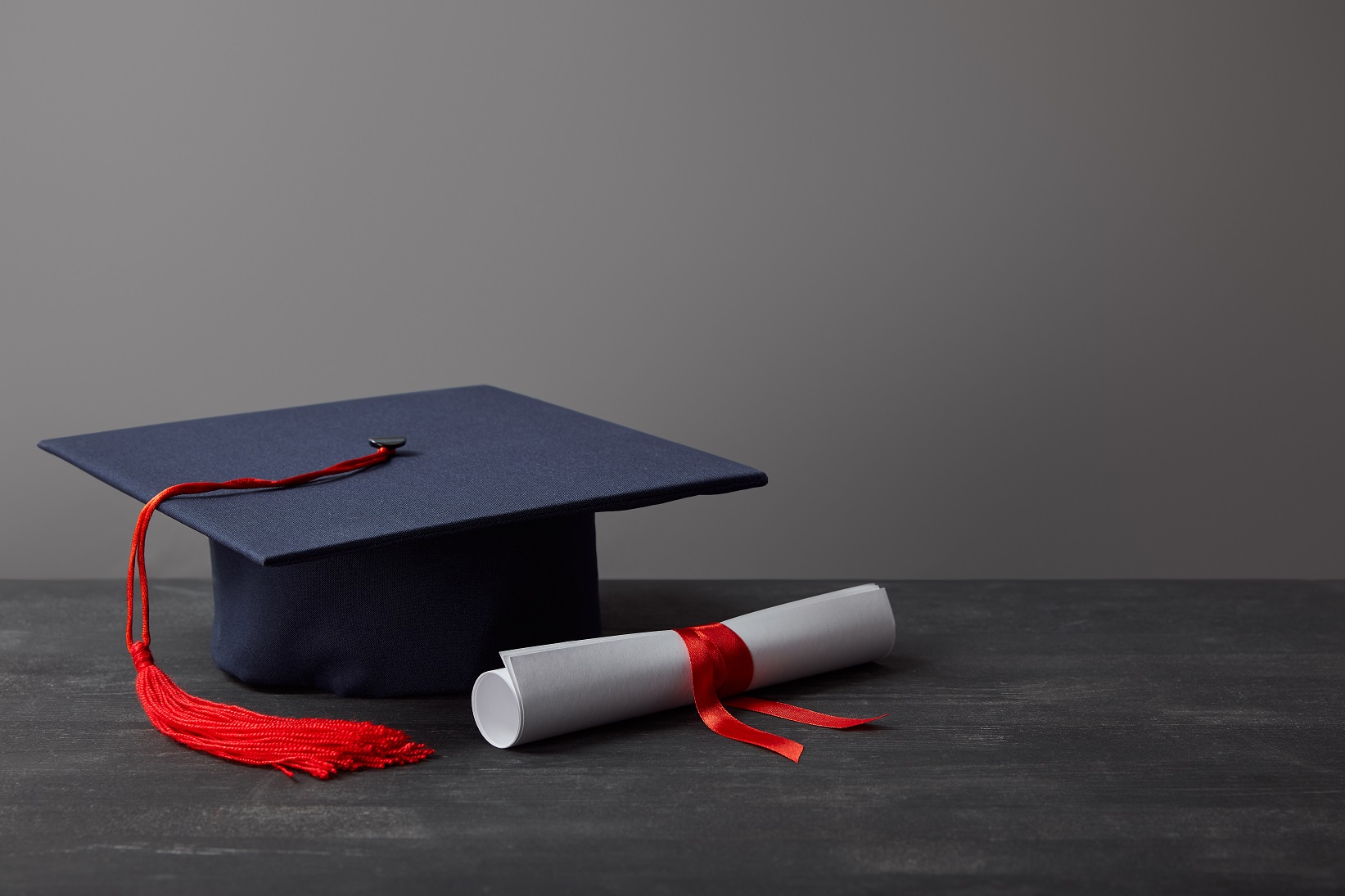 Diploma and academic cap with red tassel on dark surface on grey.